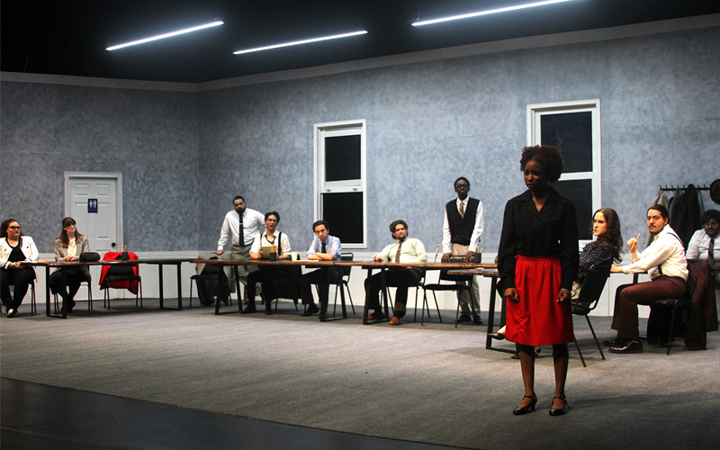 Image Description: A still from "Twelve Angry Jurors," Spring 2026. A group of people in suits look at a lone woman in a starkly lit room.