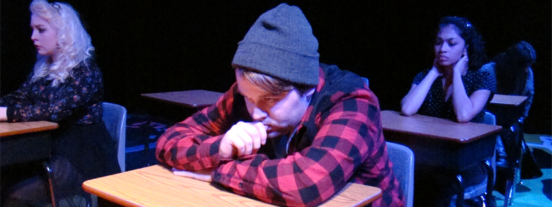 Teenager wearing a beanie and plaid shirt. He is at a school desk with his arms on the desk and chin on his arms, with an unhappy expression on his face.