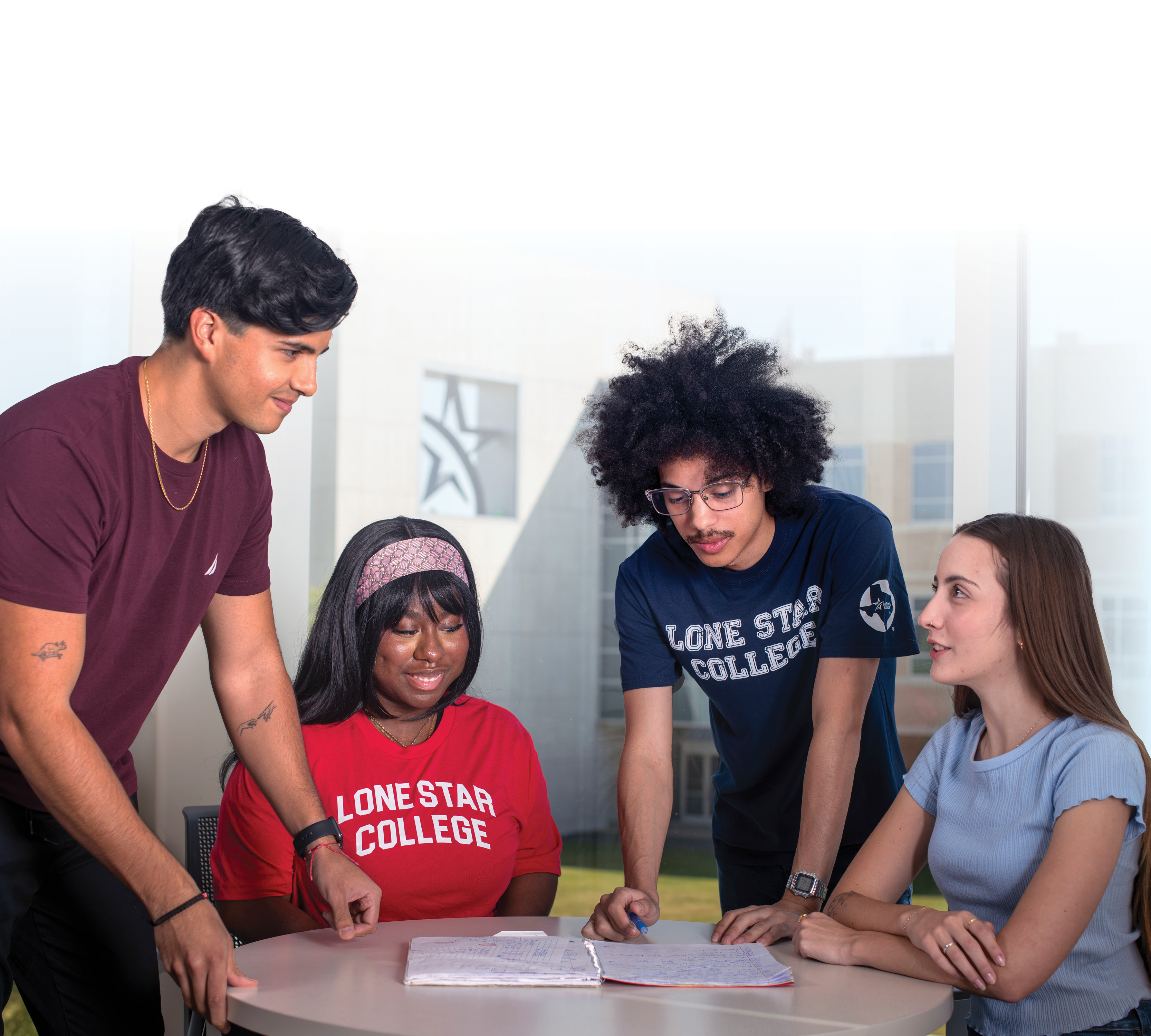 two female and two male students, surrounding a round table that has an open notebook on it, with a building on the background