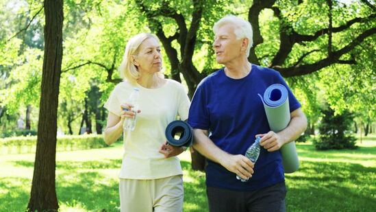 Elderly couple drinks water after outdoor exercise session in a park holding yoga mats.