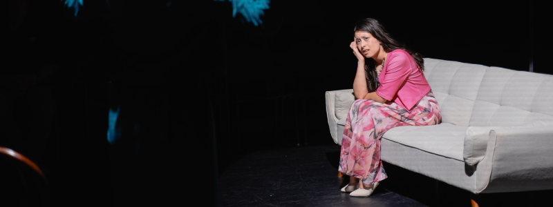 Woman wearing pink, sitting on a white couch in a dark room, with a distressed look on her face. There is a shadowy figure in the foreground.