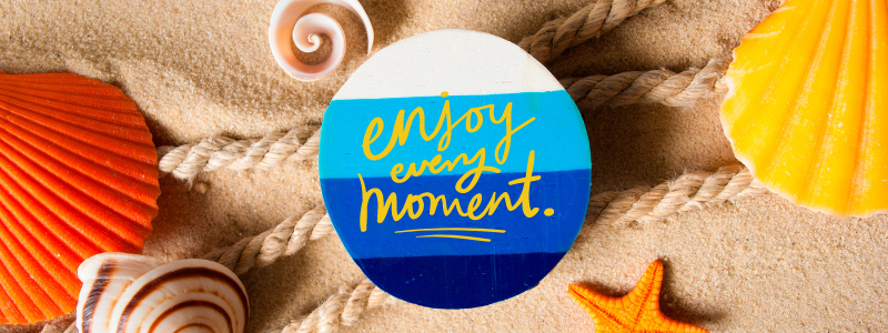 A photo with a half-orange seashell image on the left margin and a spiral-patterned seashell at the bottom left corner. A circular board comprised of colored stripes, with white at the top, followed by sky blue, ocean blue, and navy blue at the bottom, is inscribed with yellow text that reads "Enjoy every moment." A half-orange starfish sits at the bottom, slightly to the right corner, and a bright yellow seashell rests in the corner of the photo. All the marina objects are laid out on a sandy background, with three linen ropes on top of the sand and under the blue-striped circular board.